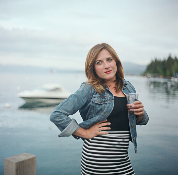A woman standing outdoors near a body of water, holding a drink, with boats in the background.