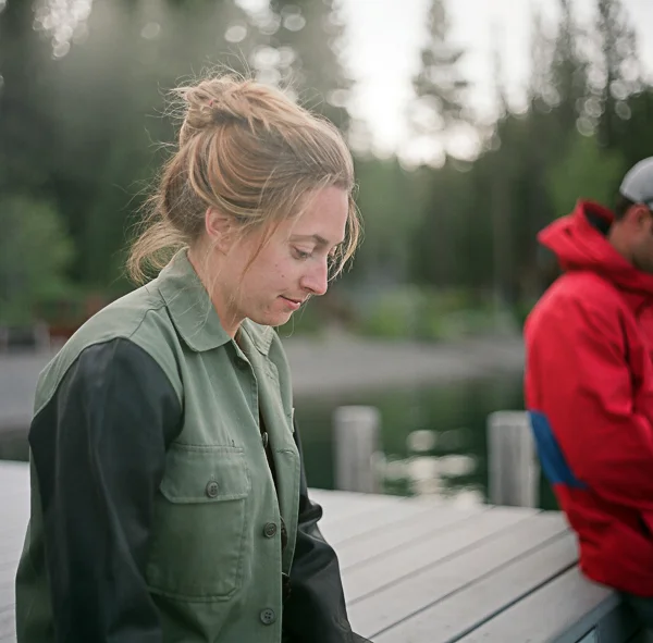 A woman with blonde hair in a bun, wearing a green jacket, standing outdoors near a body of water, with trees in the background.