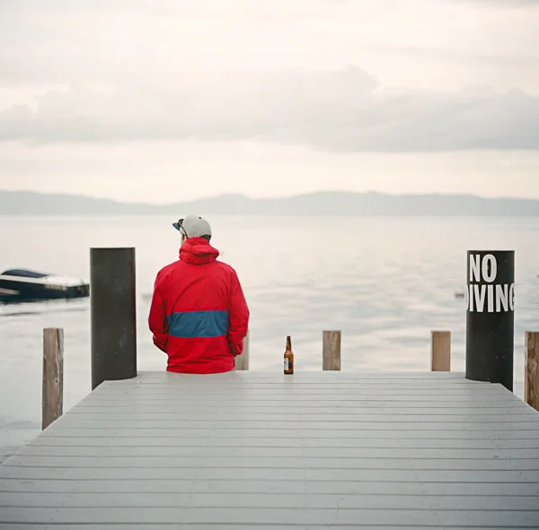 A person in a red jacket and gray cap stands on a dock looking at a body of water, with a bottle placed on the dock and a black and white "NO DIVING" sign on the right side.