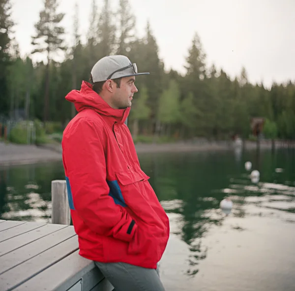 A young man in a red jacket and gray cap leaning against a wooden dock by a lake, with trees in the background.