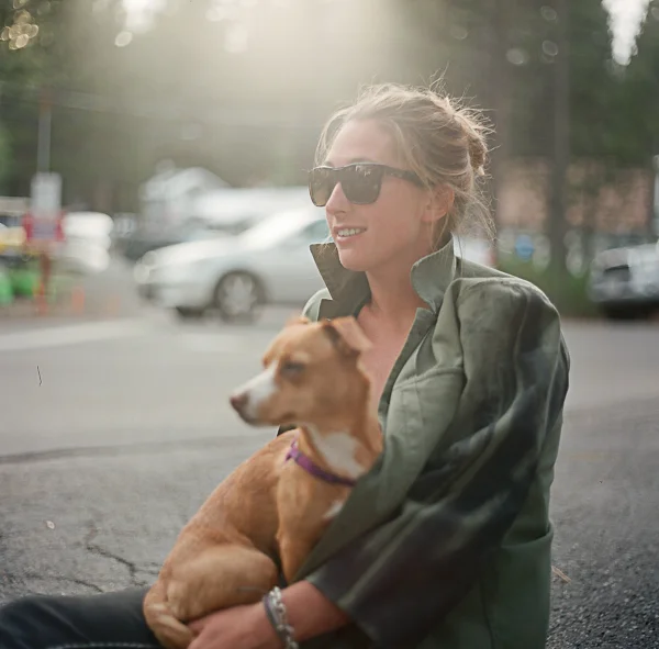 A woman wearing sunglasses and a green jacket sitting outdoors, holding a small brown dog in her lap.