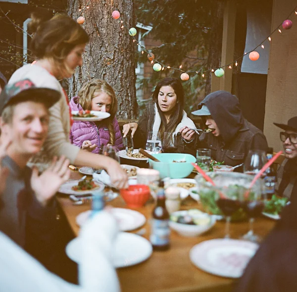 Group of people enjoying a meal outdoors at a dinner party with string lights and a large tree in the background.