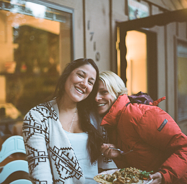Two women sitting close together at a restaurant, smiling, with a plate of food in front of them, inside a warmly lit space with large windows.