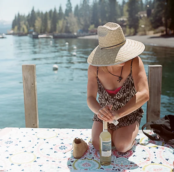 Woman kneeling on a dock by the water, wearing a large straw hat and a patterned sleeveless dress, opening a bottle of wine with a picnic bag nearby.