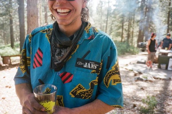 Person smiling and holding a drink outdoors in a forested area, wearing a colorful patterned shirt and a bandana.