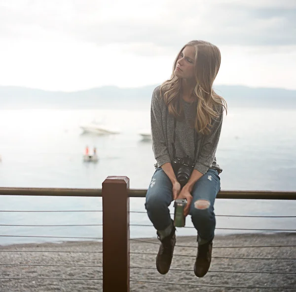 Young woman sitting on a railing near a body of water during daytime, holding a camera with a lens and a small green object, with boats and distant mountains in the background.