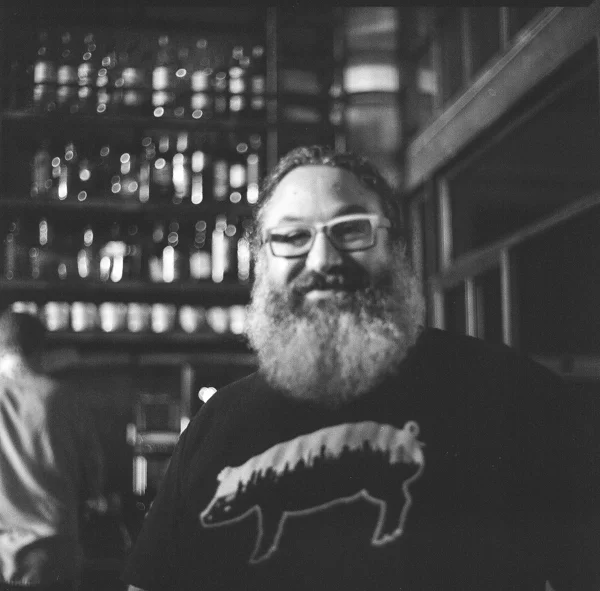A man with glasses and a full beard smiling, standing in a bar or restaurant with a background of bottles on shelves.
