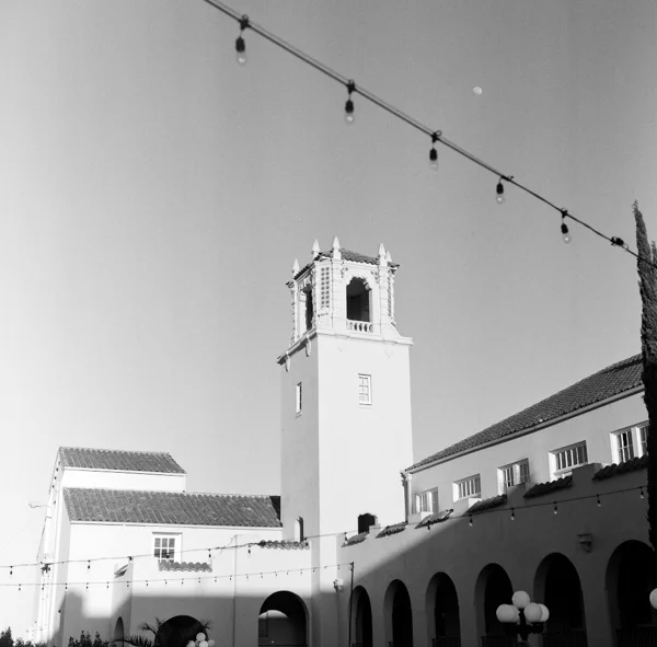 Photo of a tall clock tower with a decorative top against a sky with a visible moon, shot in black and white. Surrounding buildings with arched walkways and string lights are also visible.