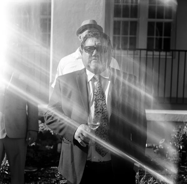 A man in a suit and sunglasses holding a glass of wine, standing outdoors with sunlight streaming in.