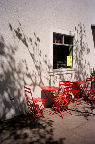 Red outdoor tables and chairs outside a building with a window and shadows cast by nearby trees.