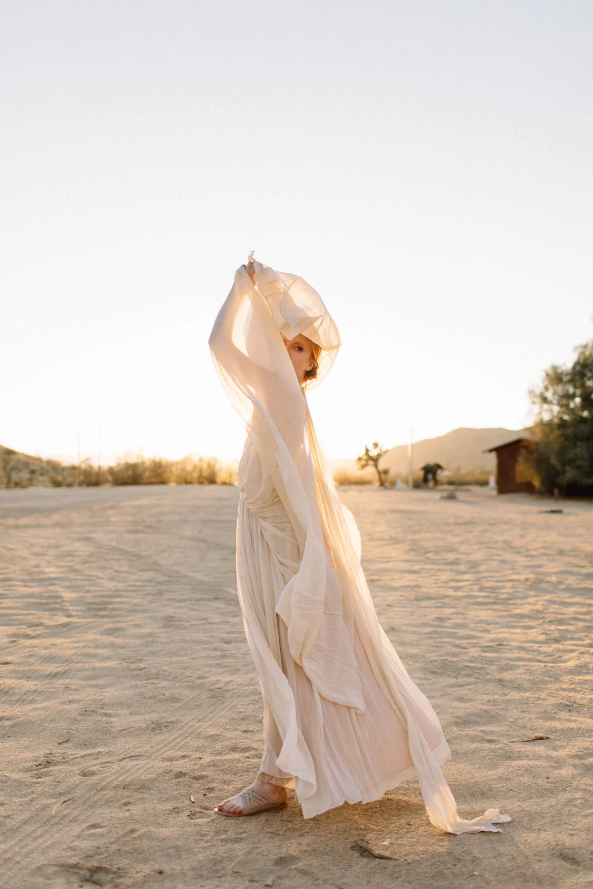 Woman in a flowing beige dress and sandals standing in a desert at sunset, partially covering her face with a sheer veil.