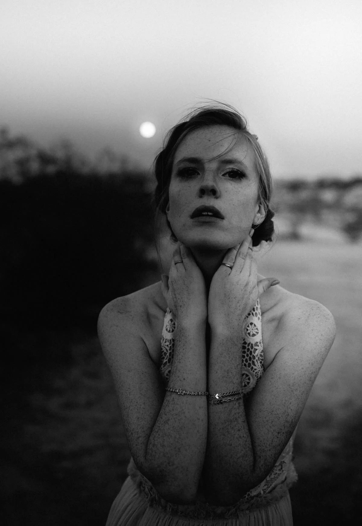 Black and white portrait of a woman with short hair, freckles, and a lace top, standing outdoors at dusk with the moon in the background.