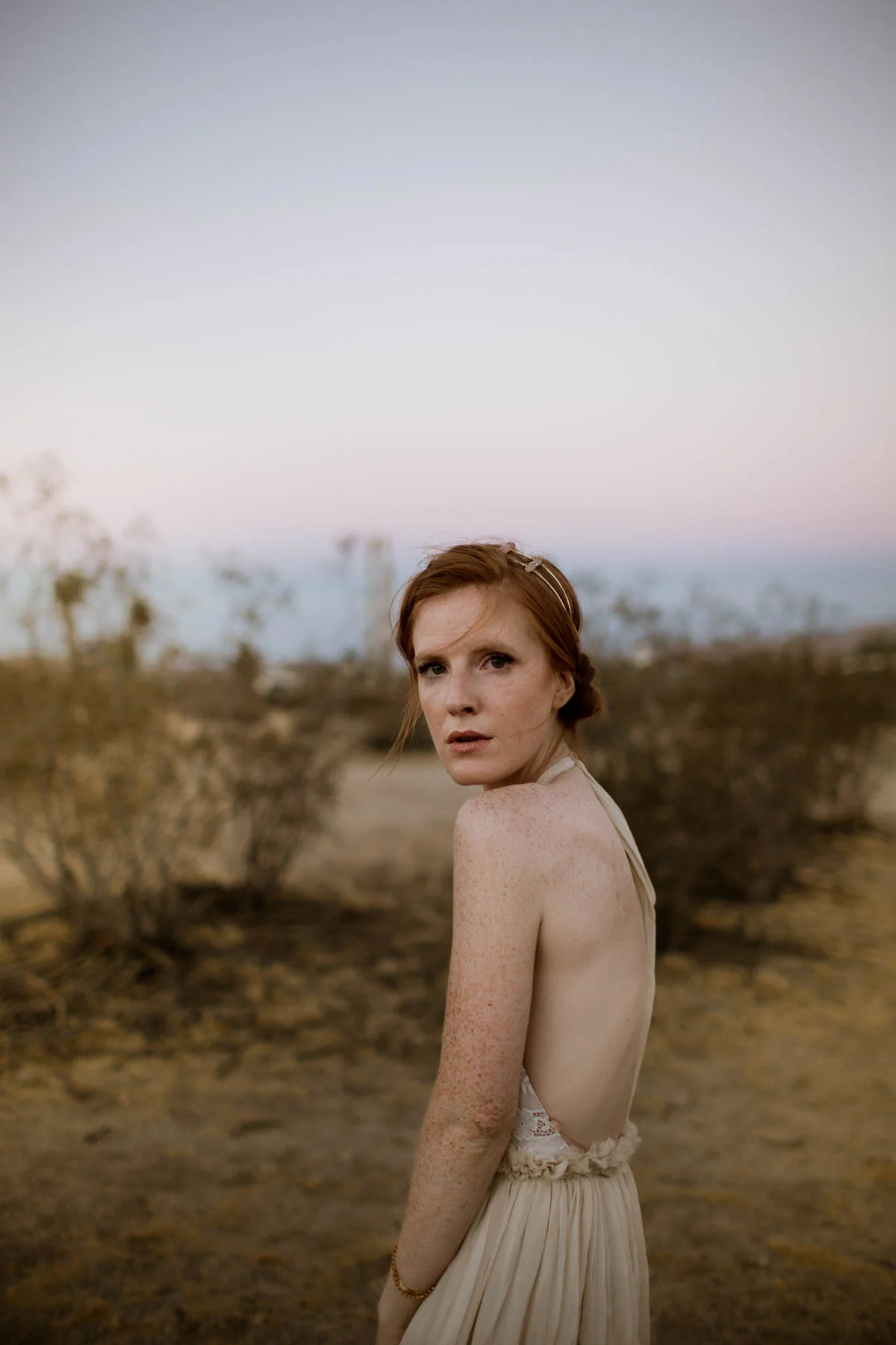 A young woman with red hair, freckles, and a light-colored dress standing outdoors in a desert landscape at dusk, looking over her shoulder.