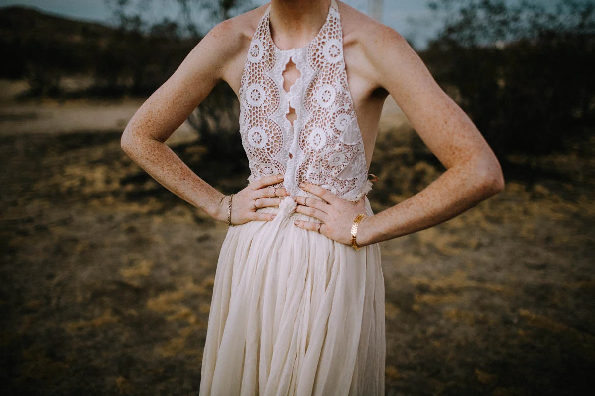 A woman wearing a white lace top and a cream-colored skirt, standing outdoors with hands on hips.