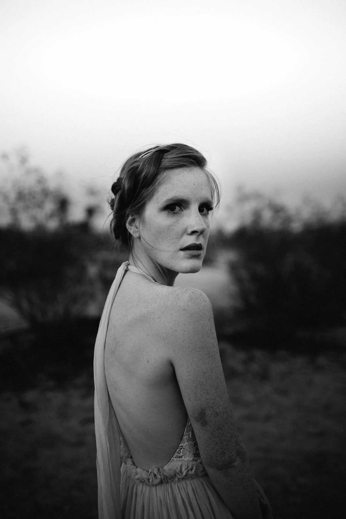 A black and white photo of a young woman with freckles and a braided hairstyle, looking over her shoulder outdoors.