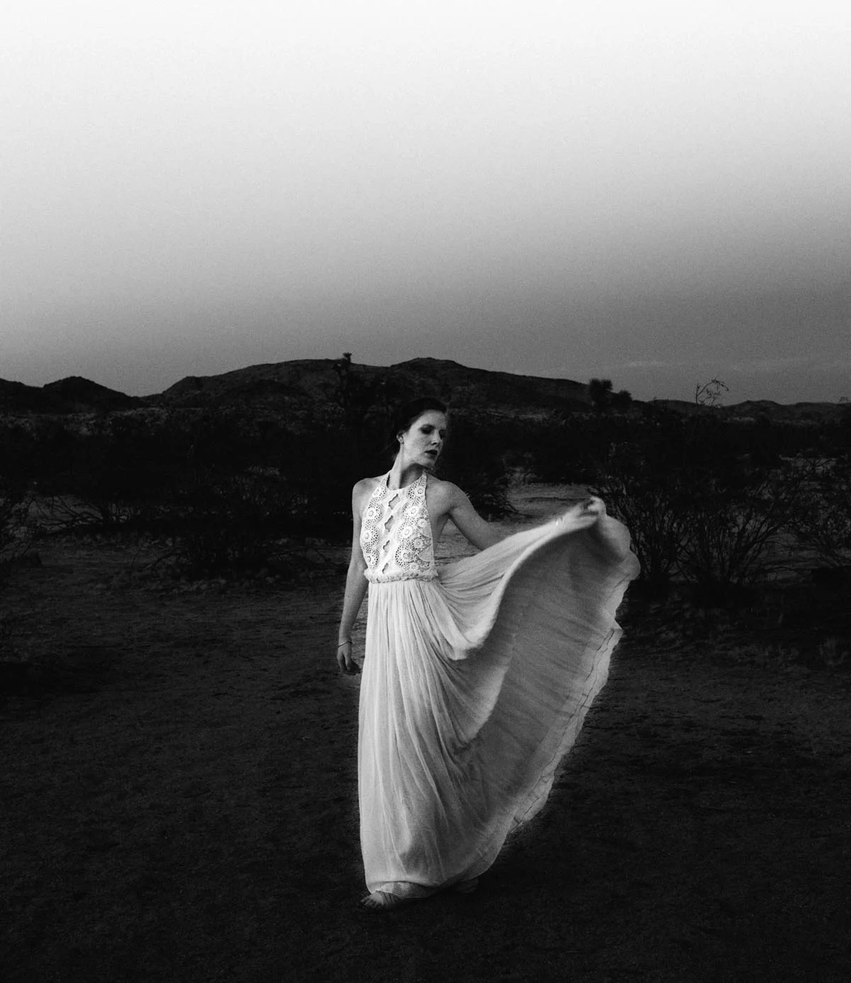 A woman in a long flowing dress standing in a desert landscape during twilight, holding part of her dress and looking to the side.