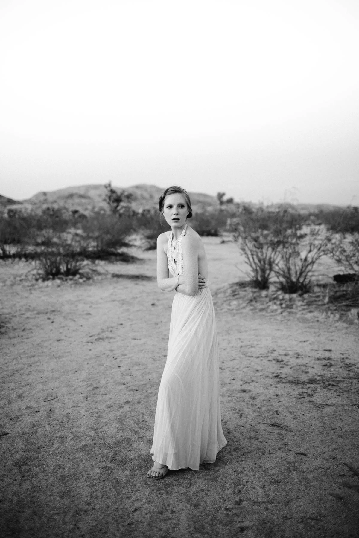 A young woman in a vintage style dress stands alone in a desert landscape with sparse bushes and distant hills, looking to her left with a serious expression in black and white.