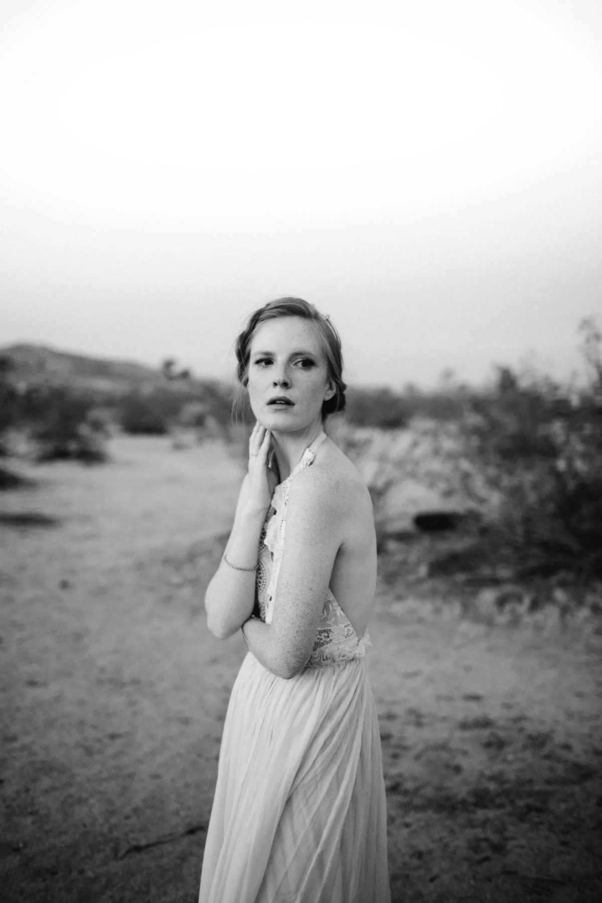 A black and white photo of a woman in a long dress standing outdoors in a desert landscape with hills and sparse vegetation in the background.