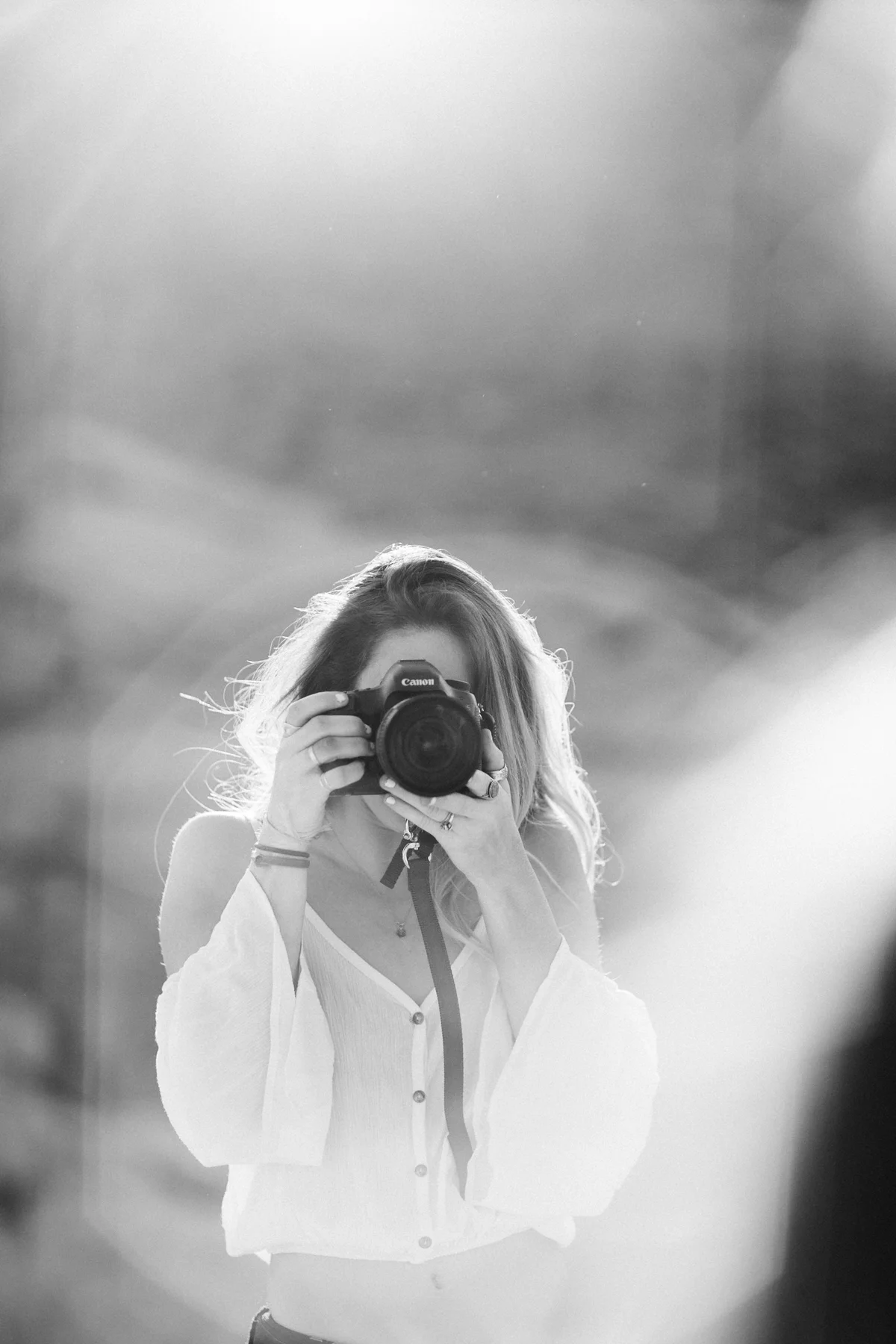 Black-and-white photo of a woman taking a picture with a Canon camera, her face obscured by the camera, wearing a white blouse, with long hair, in front of a mirror.