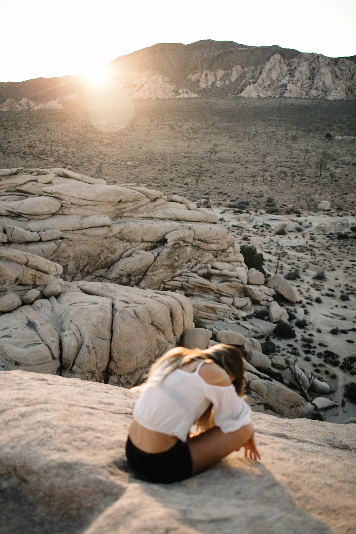 A woman in a white top and black shorts is sitting on a rocky desert landscape, with a mountain range and the setting sun in the background.