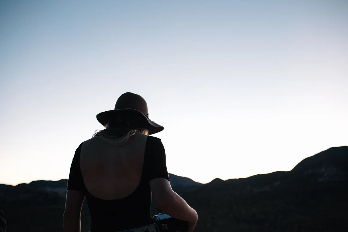 Silhouette of a woman wearing a hat and a black outfit, standing outdoors during sunset or sunrise, with mountains in the background.