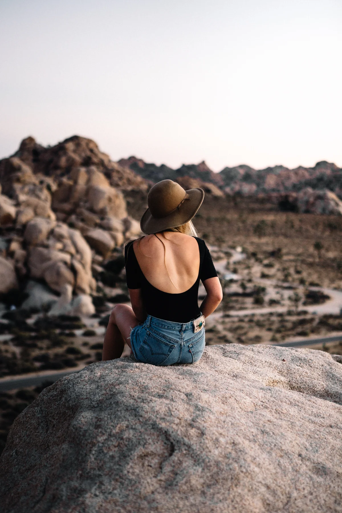 A woman sitting on a large rock overlooking a desert landscape at sunset, wearing a large sun hat, black open-back top, and denim shorts.
