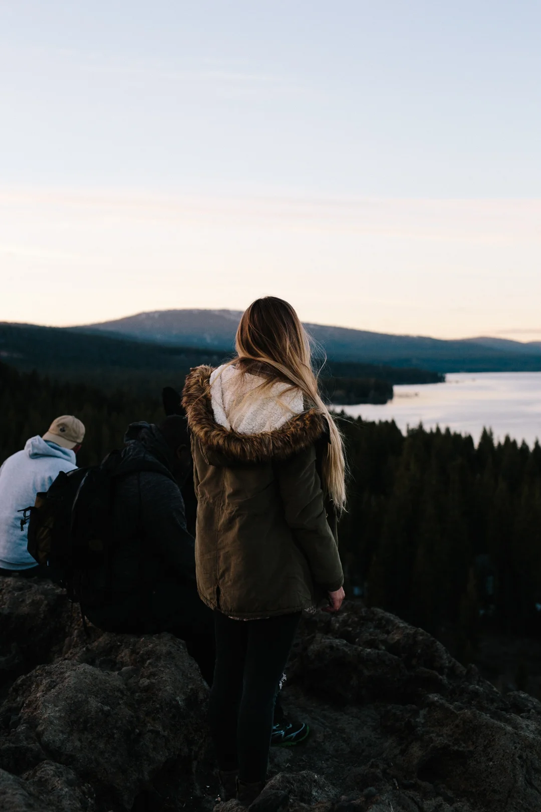 A young woman with long blond hair in a brown coat with a fur-lined hood standing on rocky terrain, overlooking a scenic view of a lake and forested mountains at dusk, with two seated men nearby.
