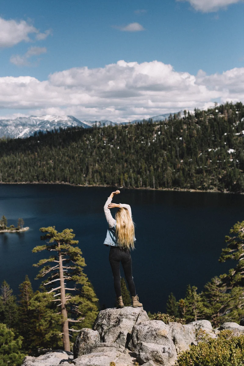 A woman standing on a large rock overlooking a lake surrounded by pine trees and mountains with snow, under a partly cloudy sky.