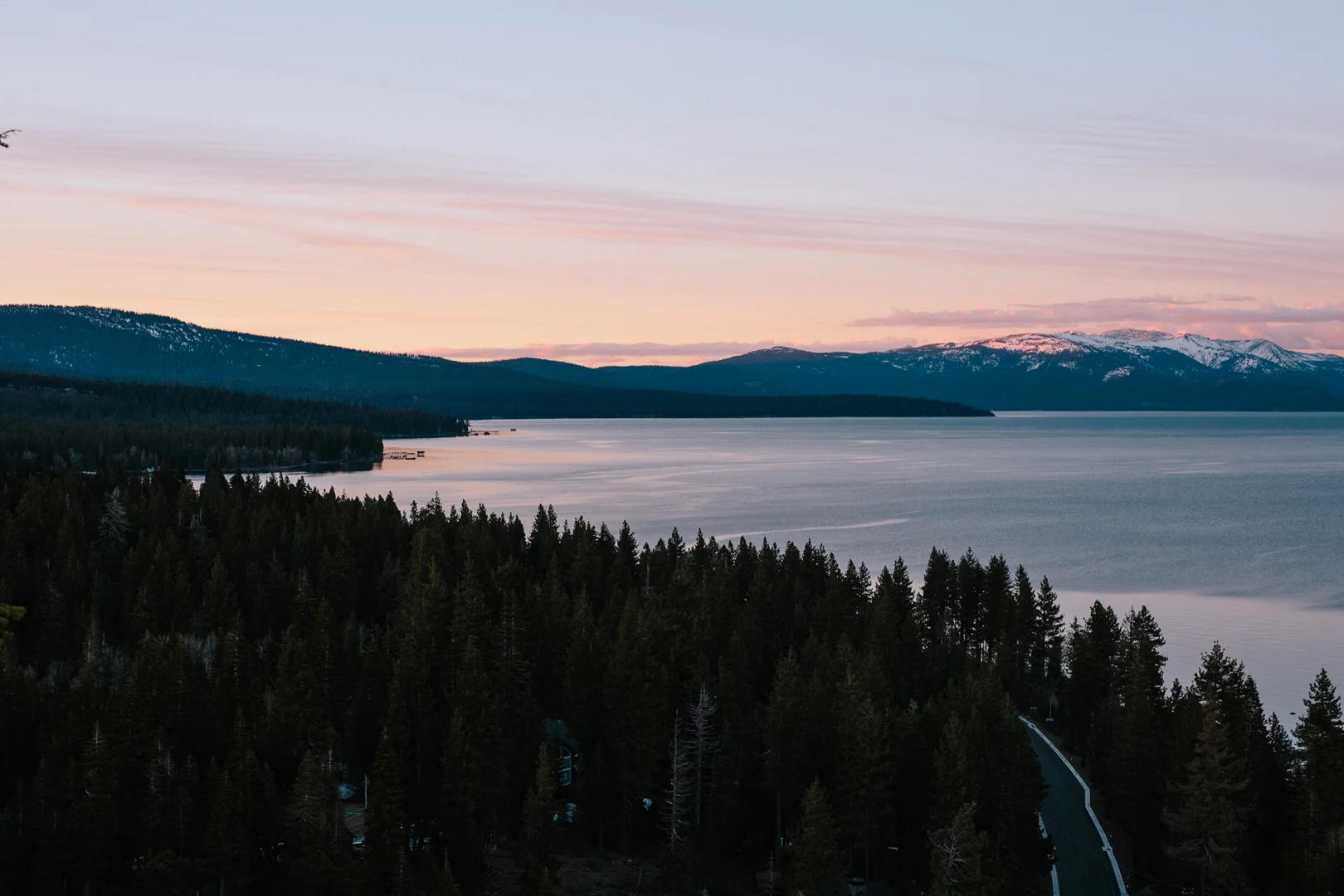 Scenic view of a large lake surrounded by pine trees and snow-capped mountains, with a pastel-colored sky at sunset.
