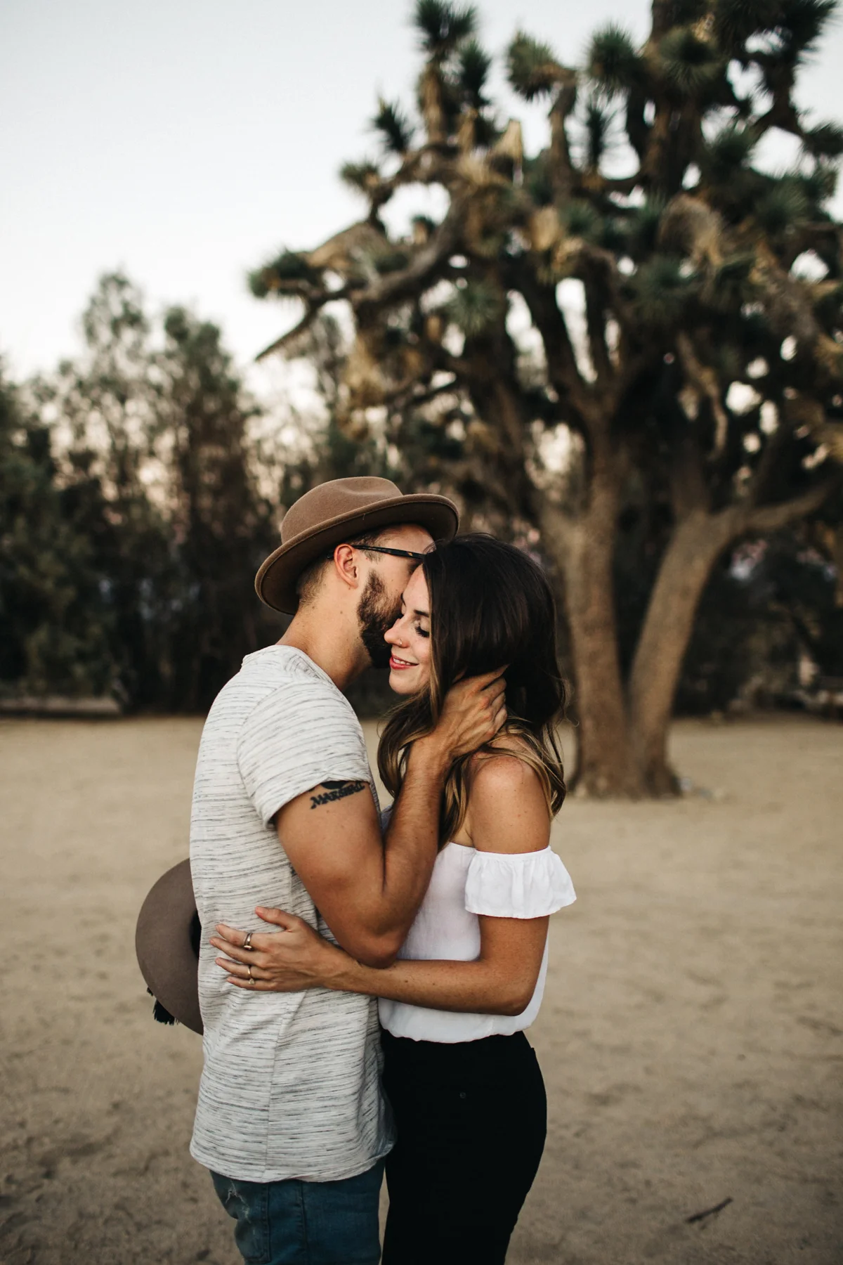 A couple in love embracing and smiling in front of a large tree in an outdoor setting during sunset.
