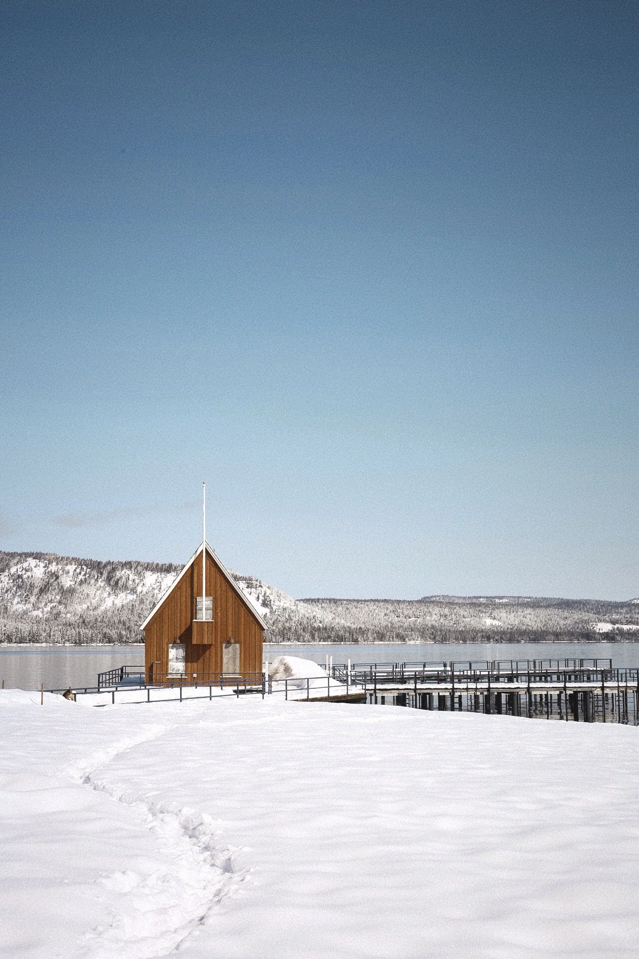 A wooden chapel on a snowy landscape beside a lake with snow-covered mountains in the background and a clear blue sky.