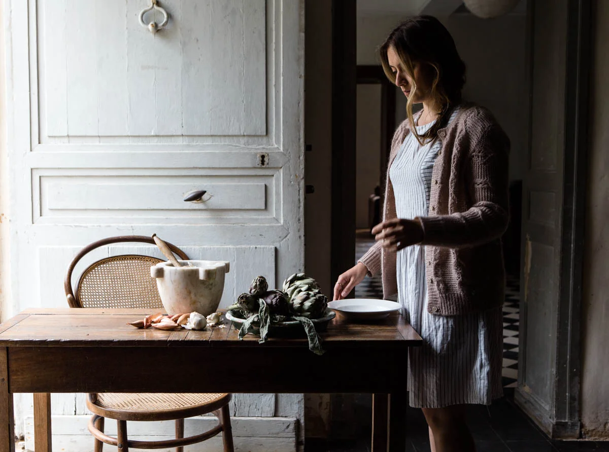A woman in a brown cardigan and striped dress standing near a wooden dining table with artichokes, garlic, and a mortar and pestle. The room has vintage decor with white paneled walls and checkered floor.