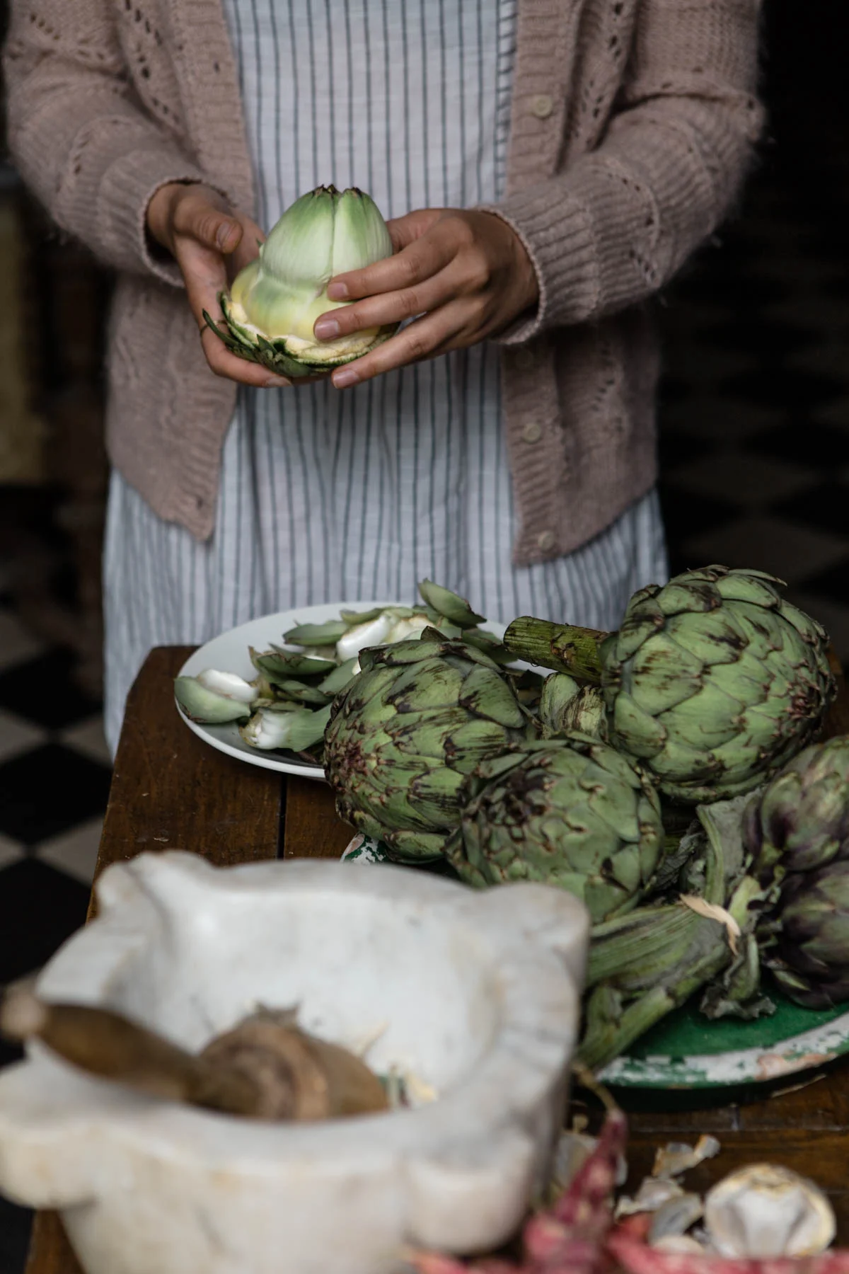 Person holding an artichoke over a table with more artichokes, garlic, and other vegetables.