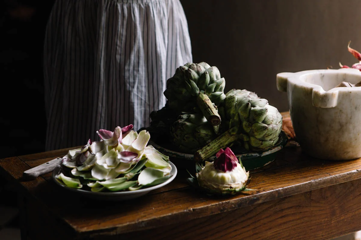 Fresh artichokes and flower petals on a wooden table with a mortar and pestle in the background.