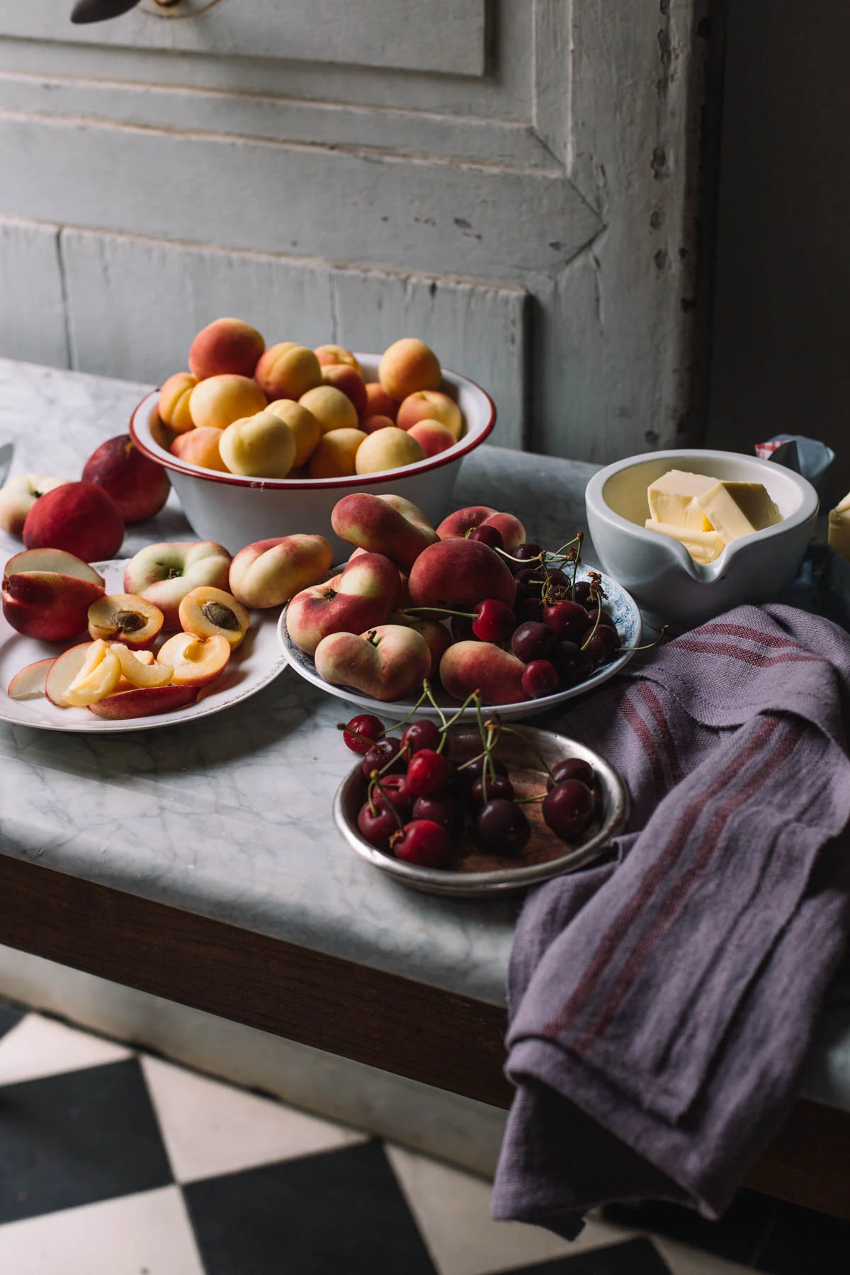 Various fresh peaches, nectarines, and cherries arranged on plates and bowls on a marble table, with a bowl of butter and a gray-striped cloth nearby.