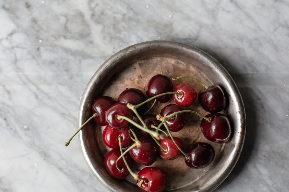 A small round metal bowl filled with red and dark purple cherries on a gray marble surface.