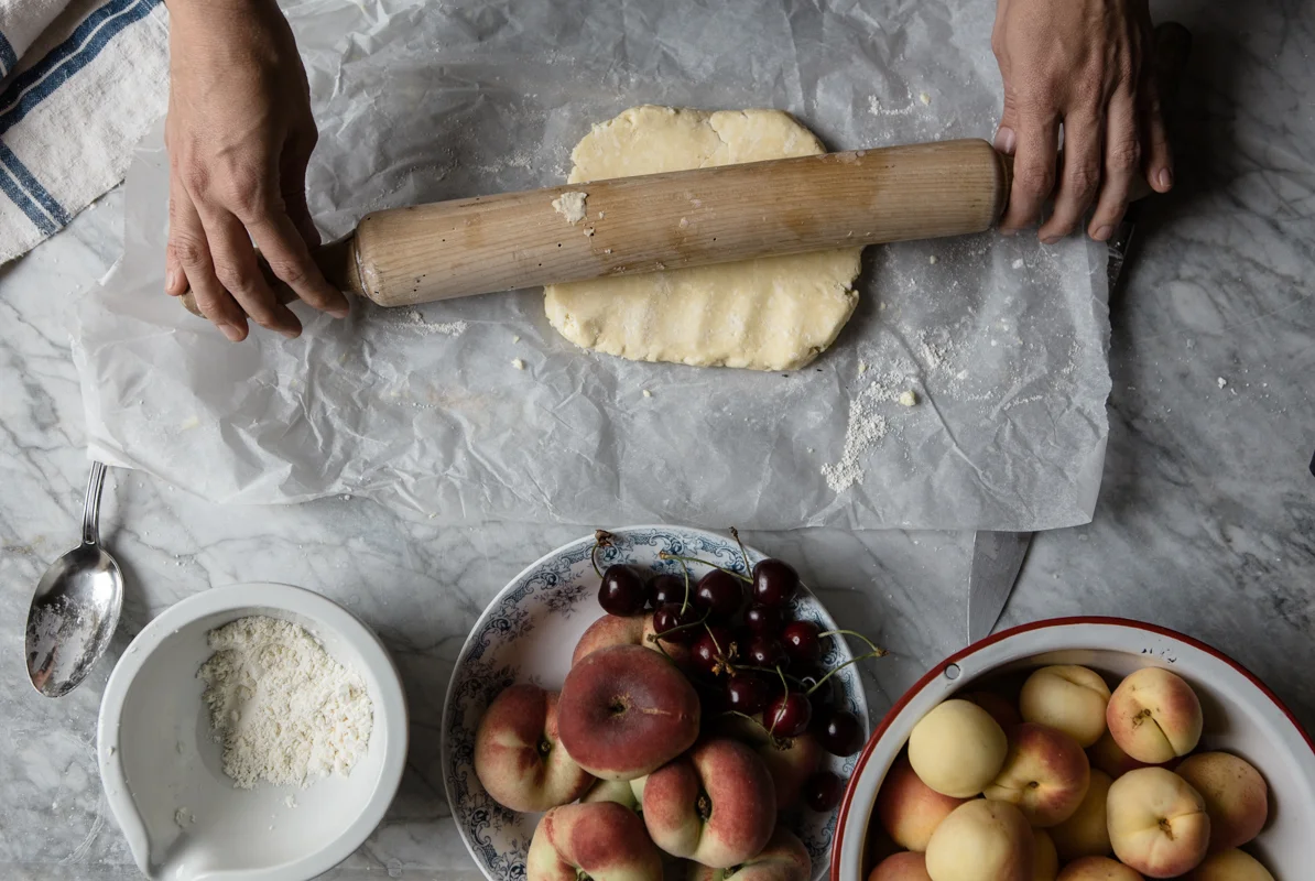Hands rolling out dough with a wooden rolling pin on parchment paper, surrounded by bowls of cherries and peaches, with a spoon and grated cheese nearby.