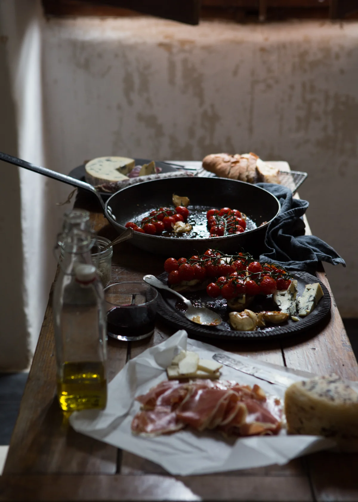 A rustic wooden table set with fresh cherry tomatoes, garlic, cheese, sliced meats, a bottle of olive oil, a glass of red wine, and a frying pan with cooked cherry tomatoes.