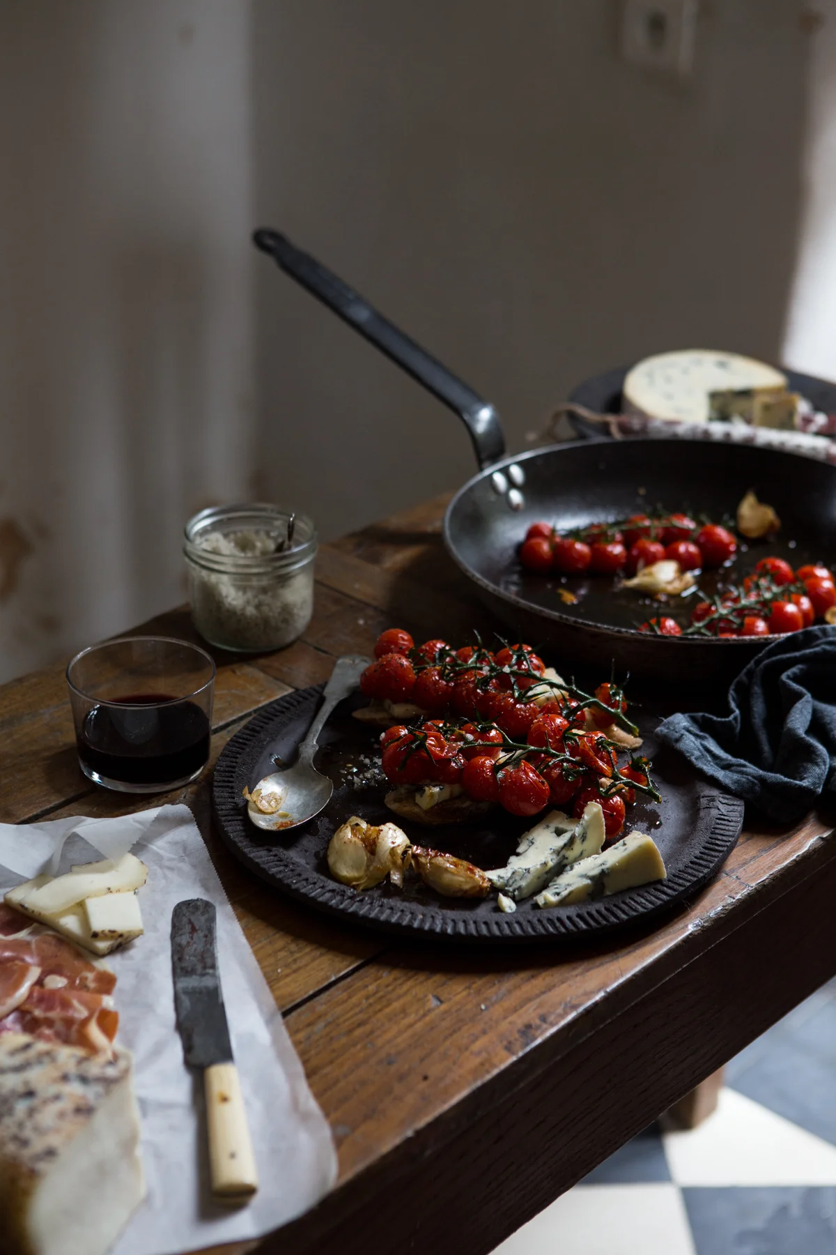 A rustic wooden table with a charcuterie board featuring cherry tomatoes on the vine, cheese, and grilled garlic. A half-empty glass of red wine, a jar of grated cheese, and a cast iron skillet with more cherry tomatoes and garlic are also on the tab