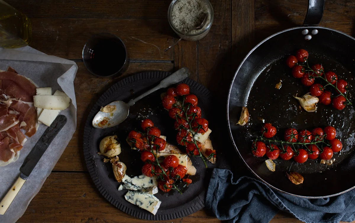 A rustic wooden table with a cast iron skillet filled with roasted cherry tomatoes and garlic, a black plate with roasted tomatoes, cheese, and bread, a glass of red wine, a jar of grated cheese, and a knife with cheese on parchment paper.