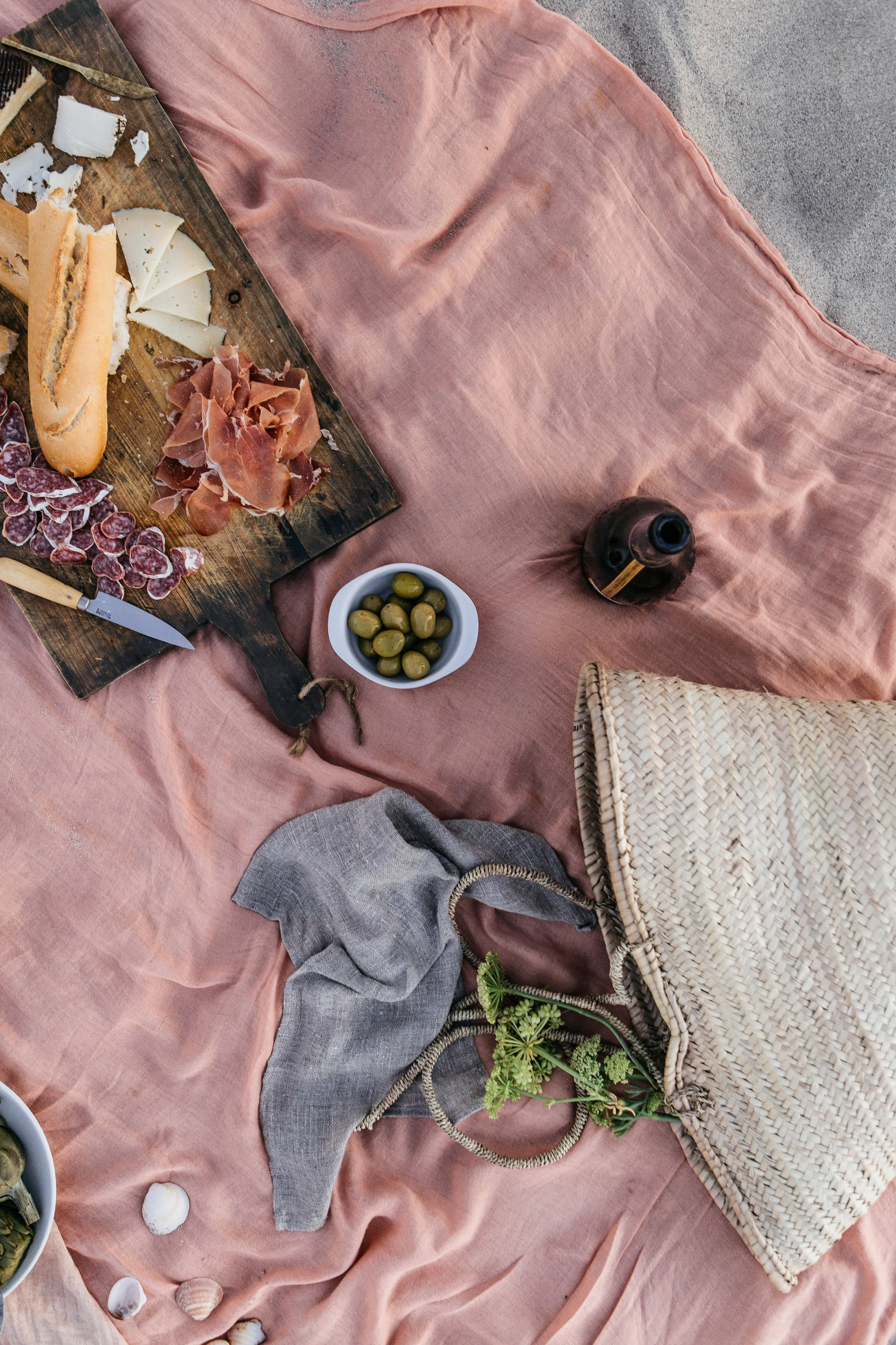 A picnic setup with a pink cloth on the ground, featuring a wooden board with cheeses and cured meats, a bowl of green olives, a wicker bag with greenery inside, seashells, and a dark bottle, suggesting a relaxed outdoor meal.