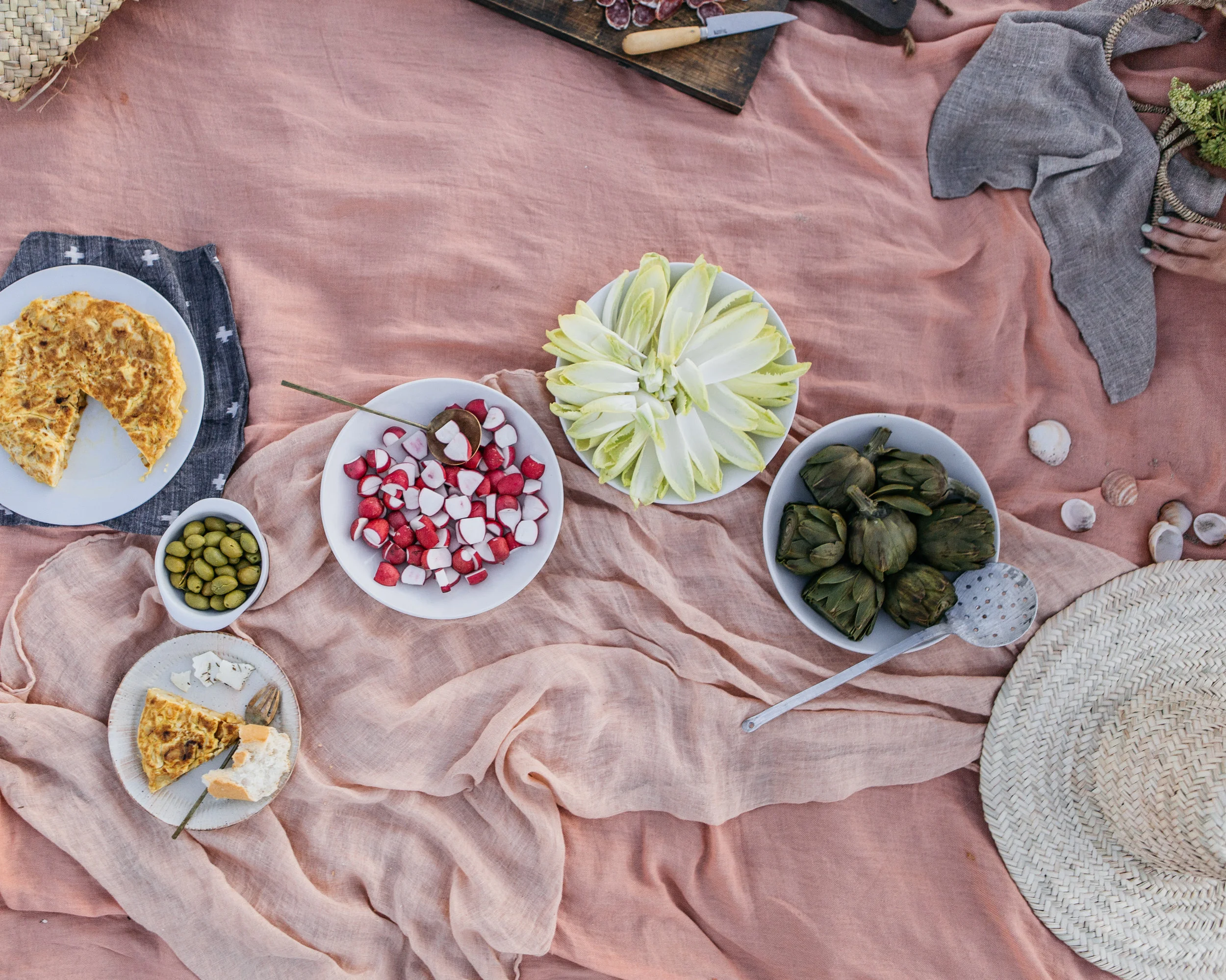 A picnic setup with various dishes including a slice of quiche on a small plate, a bowl of green olives, a plate of sliced radishes, a bowl of artichokes, and a large bunch of white-tipped endive on a pink tablecloth, with seashells scattered around,