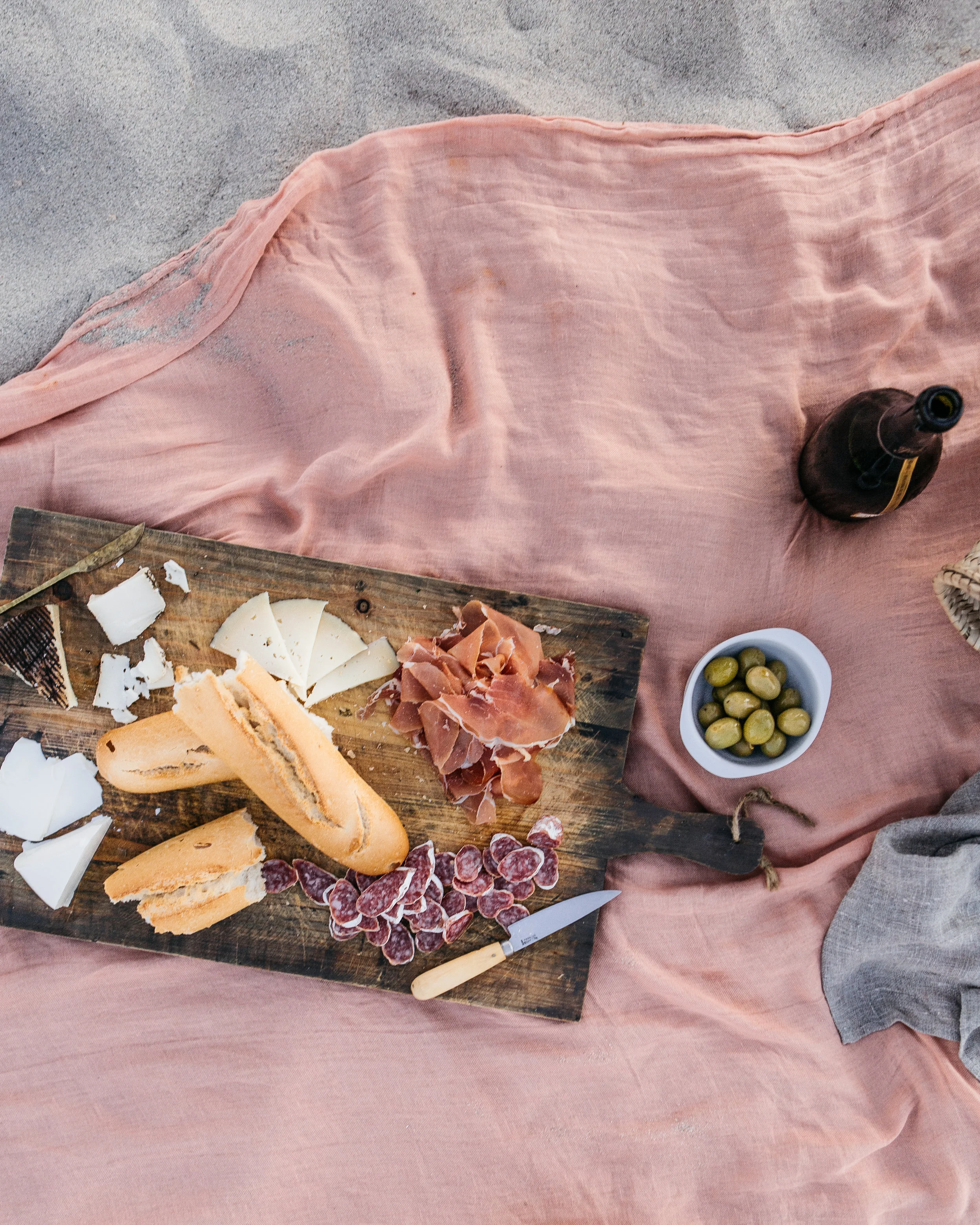 A picnic spread on a pink blanket with cheese, bread, cured meats, green olives, and a bottle of beer.