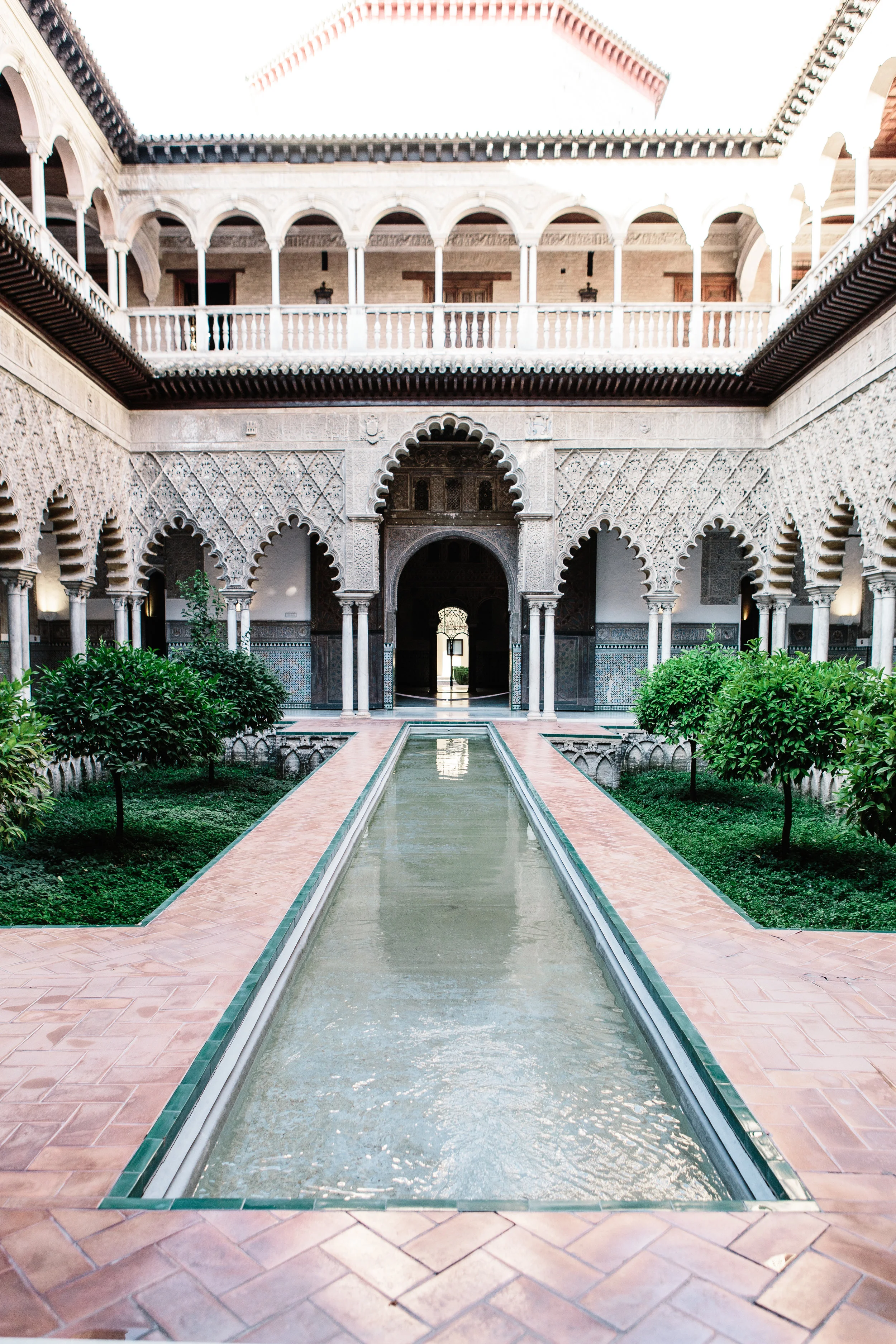 Interior courtyard of a traditional Moroccan riad with a central rectangular water fountain, surrounded by lush green plants, arched doorways, intricate stucco carvings, and a two-story gallery with columns.