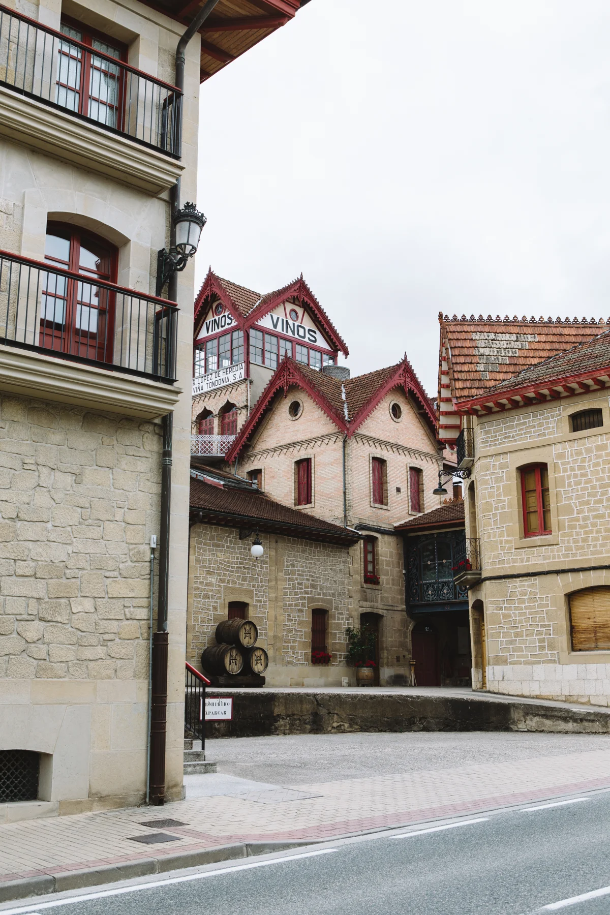 View of a European-style street with stone buildings, barrels, and a sign that says "Vinos." A building in the background has red decorative trim on the roof and windows, with signs indicating wine (Vinos).
