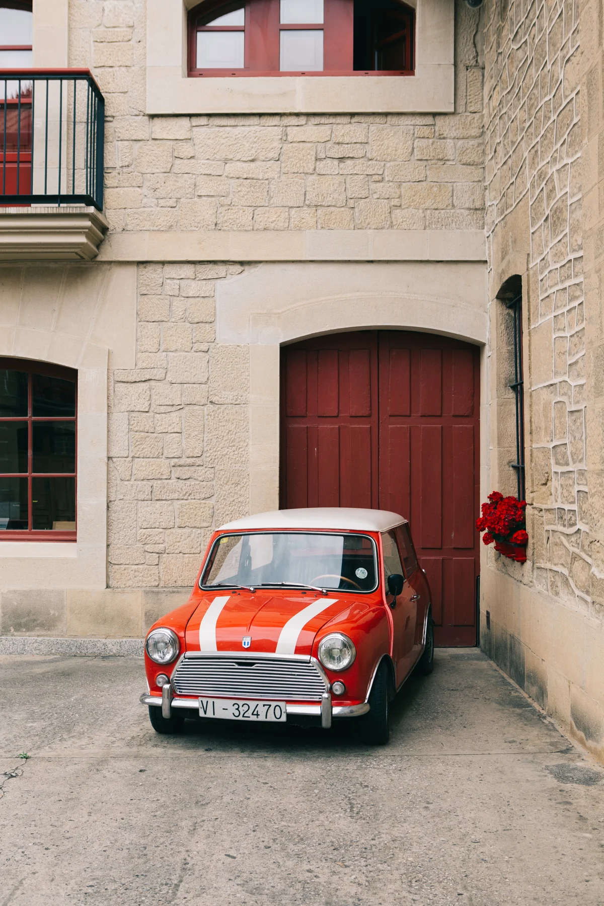A small vintage red and white car parked in front of a stone building with red wooden doors and windows, a flower box with red flowers on the wall.