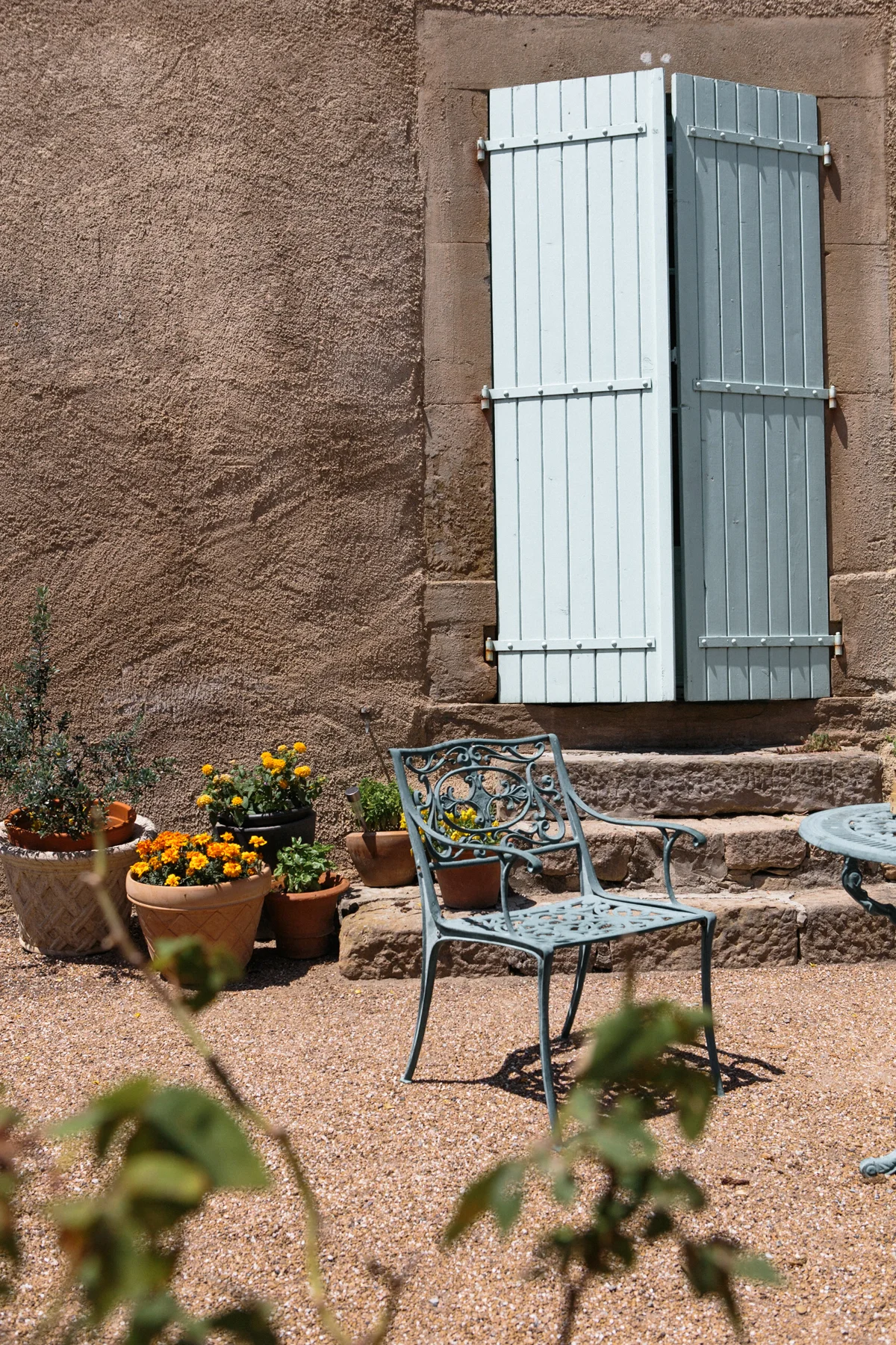 A cozy outdoor patio area with a textured brown wall, a set of pale blue shutters on the wall, an ornate metal bench, a round table, and several potted plants with colorful flowers.