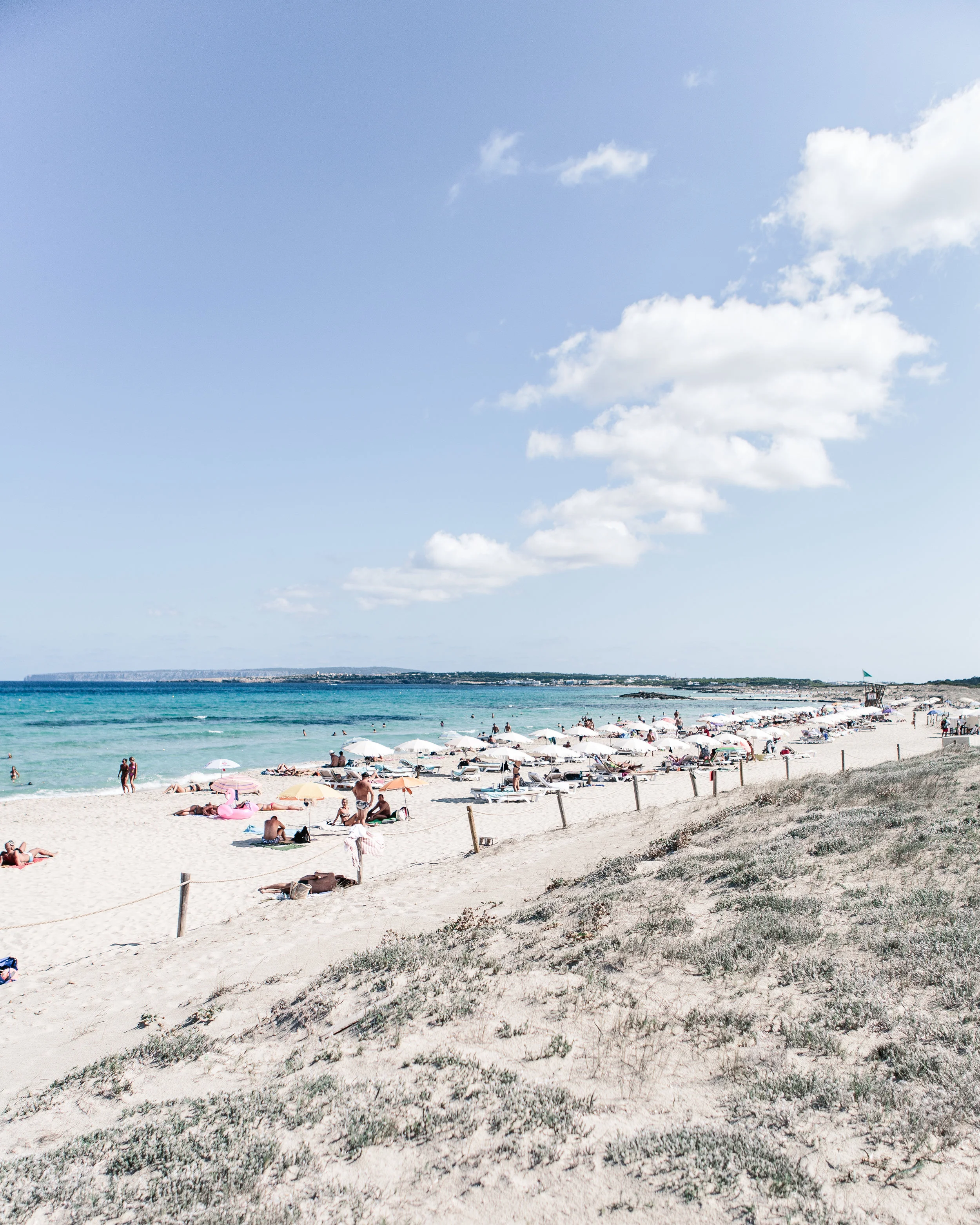Beach scene with people sunbathing and swimming under umbrellas along a sandy shoreline, with blue skies and some clouds overhead.
