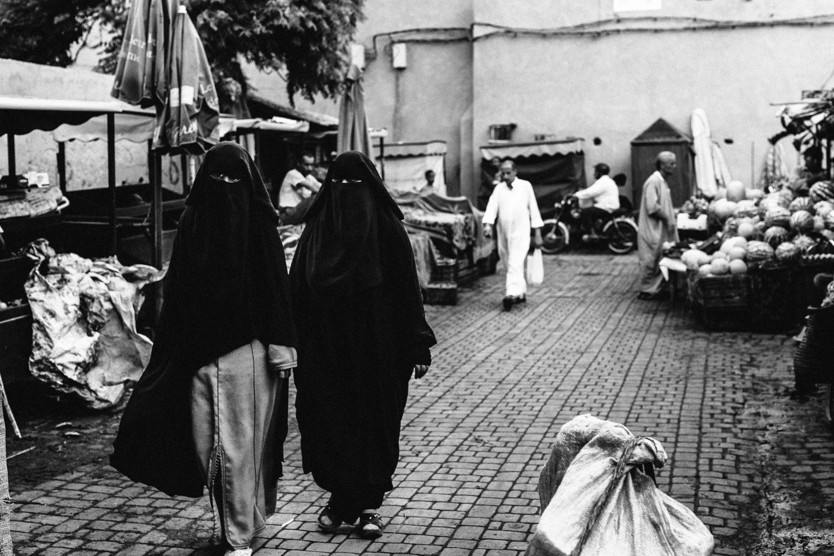Two women in black niqabs walking through an outdoor marketplace with fruit stalls, vendors, and other shoppers visible in the background.