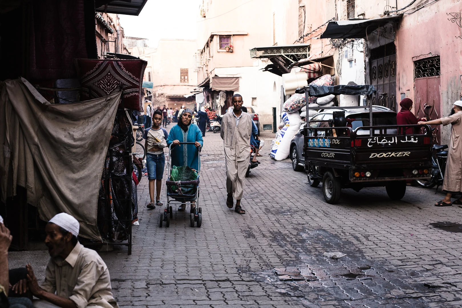 A busy street scene in an urban area with people walking, including a woman with a stroller, a man, children, and vendors; vehicles and buildings lining the street.